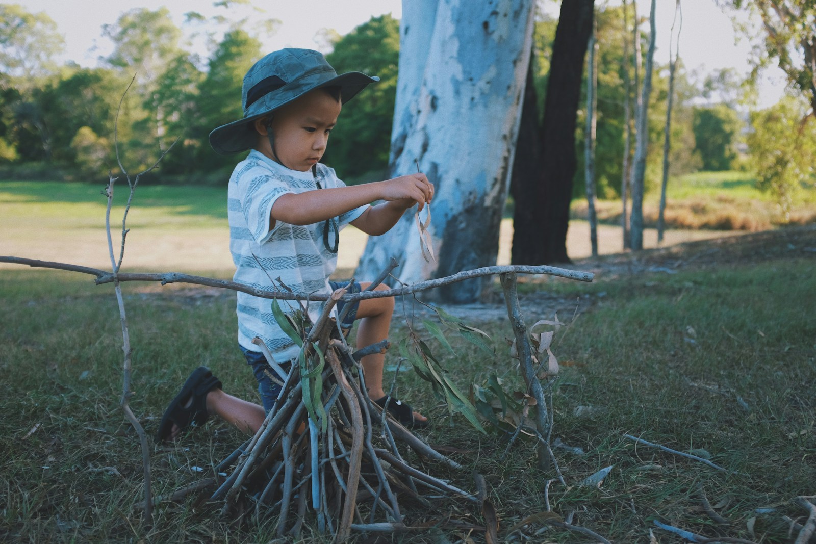 boy wearing gray hat, white-and-gray striped crew-neck t-shirt and gray shorts kneeling on ground building twigs into a fire shape during daytime
