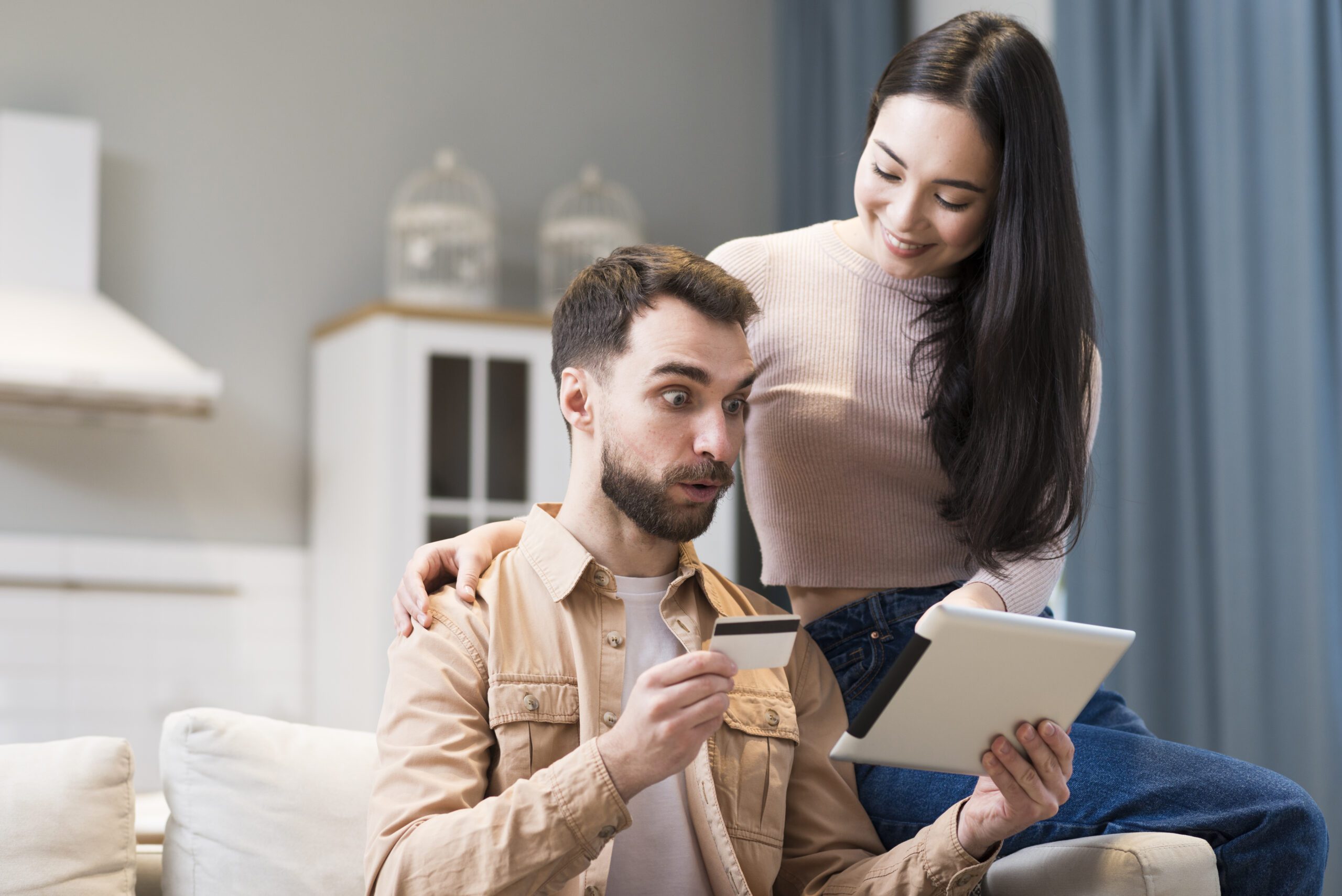 coupld sitting on sofa and looking at a tablet and the man holding a credit card and looking shocked