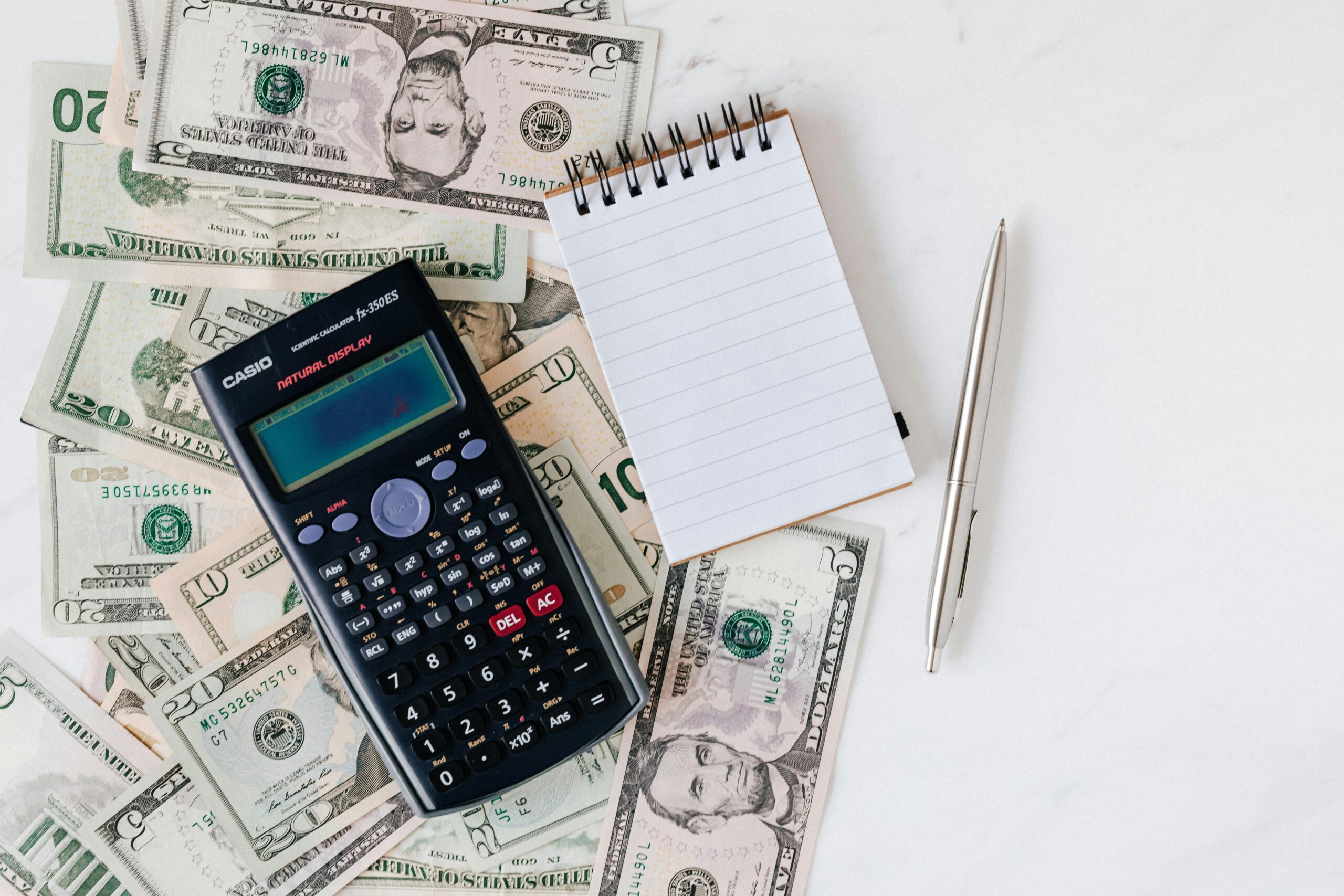 calculator, dollars, notepad and pen on white surface