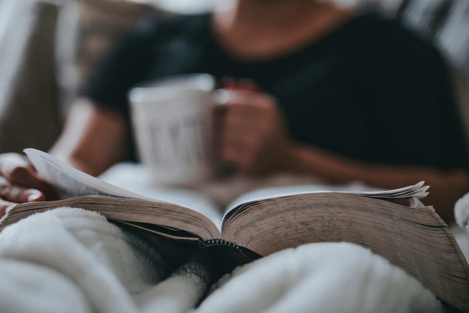 person holding coffee mug and book in bed