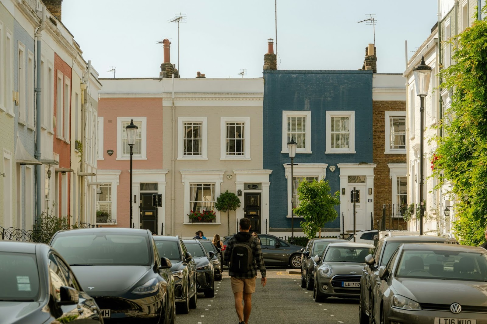 man walking down middle of street towards colourful houses at the end, with cars parked either side