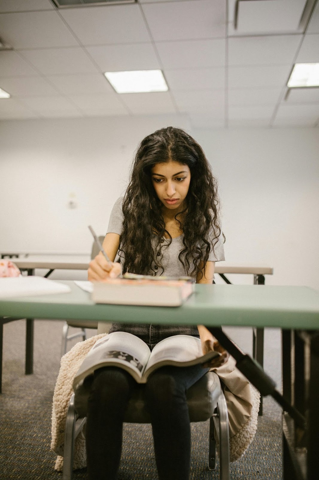 teen girl sitting at desk in classroom writing and with open book on her knee