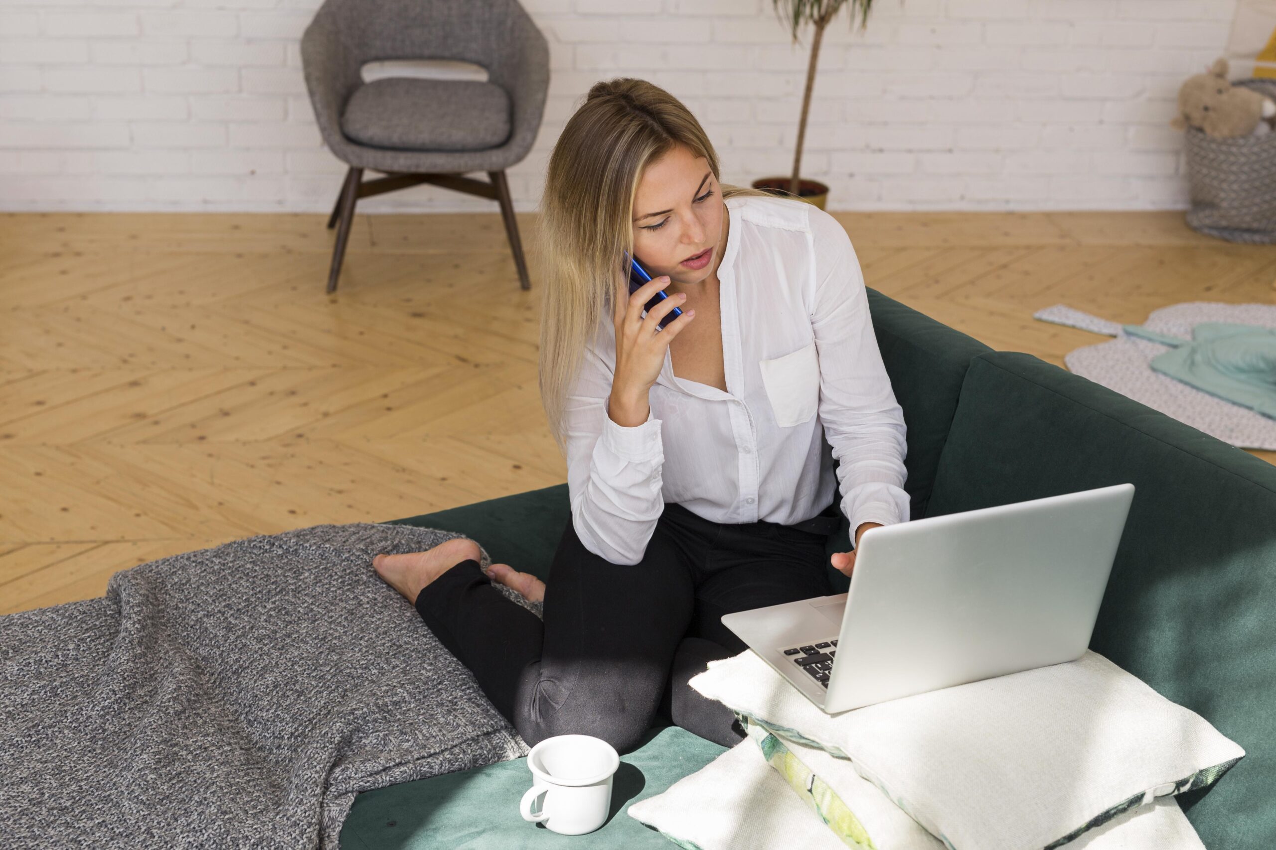 woman on phone, kneeling on sofa, with laptop in front of her, propped up on cushions