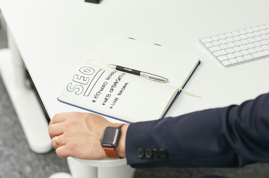 man's hand leaning on desk next to open notebook with SEO written on it and a pen on it