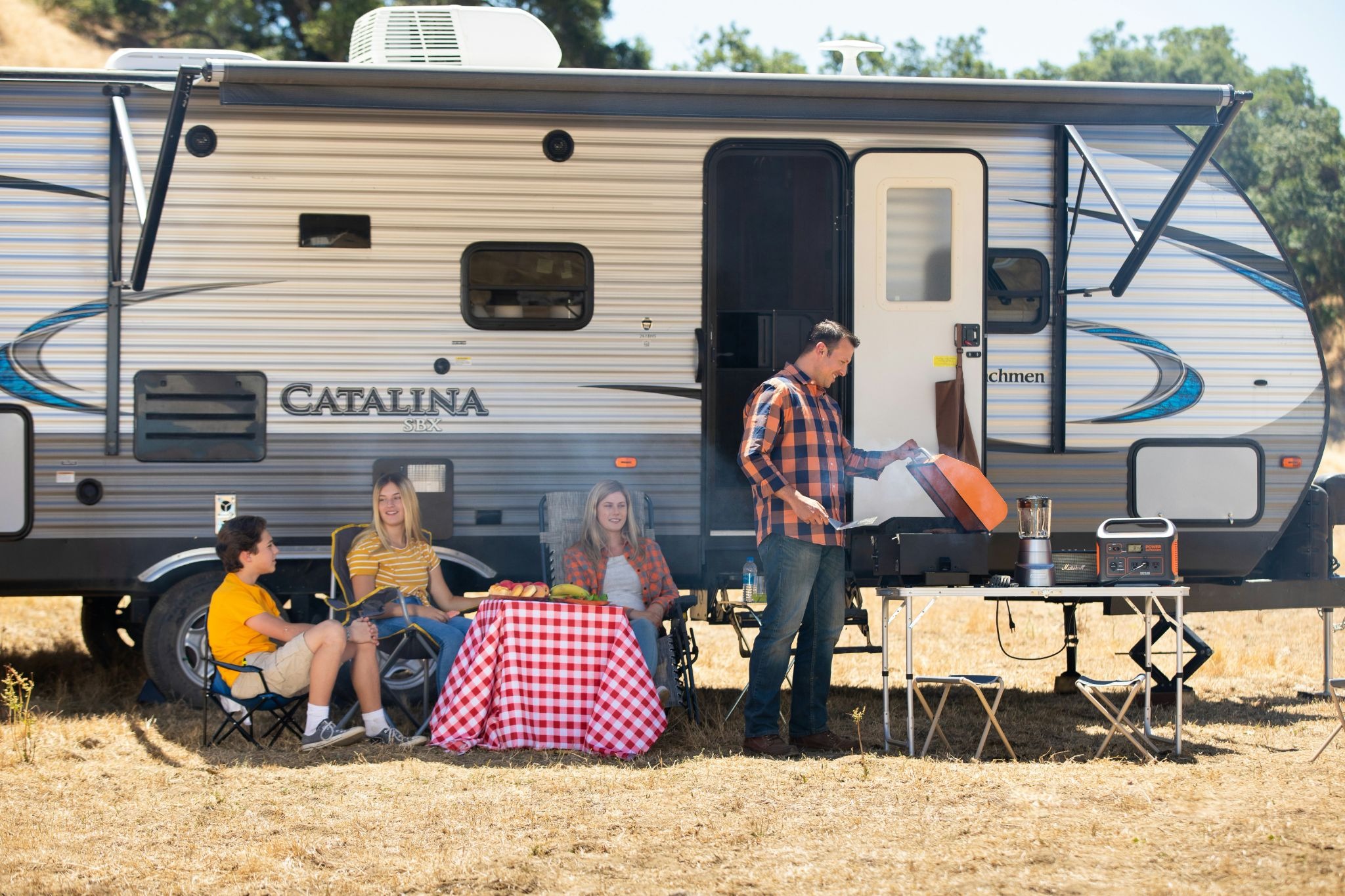 a family sitting next to an RV motorhome with the man cooking on a barbecue