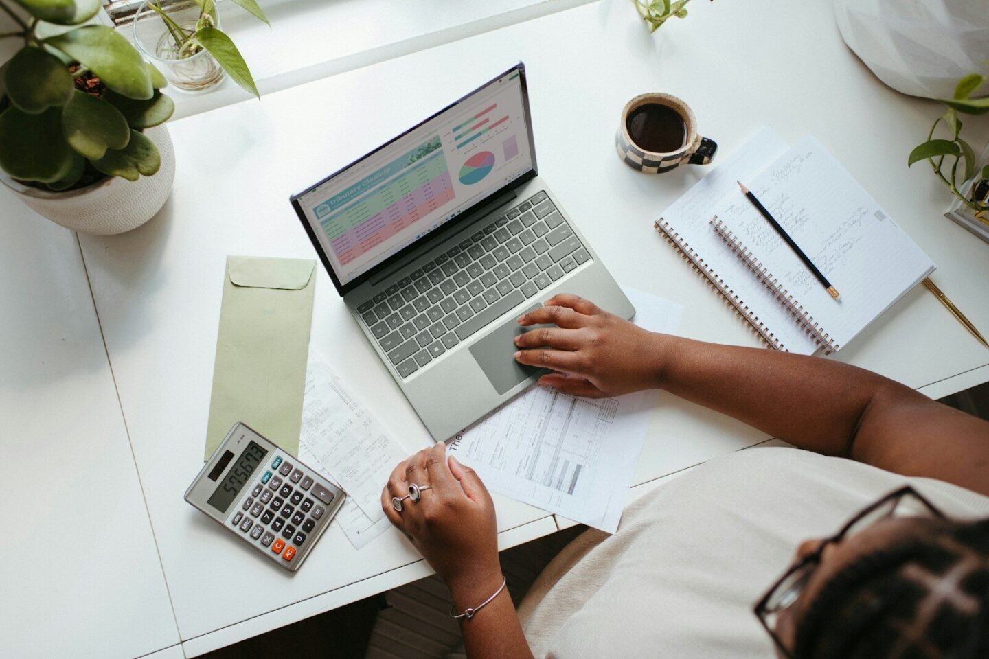 a person sitting at a table with a laptop, calculator, coffee, pens and papers