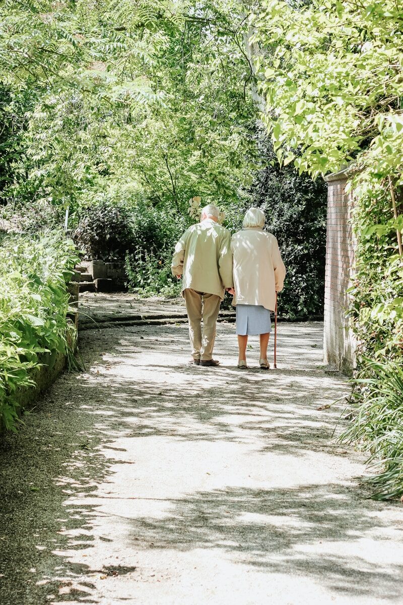 older man and woman walking on path between trees during daytime