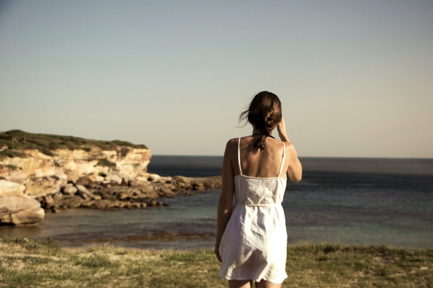 woman in white dress standing on grass  facing the sea