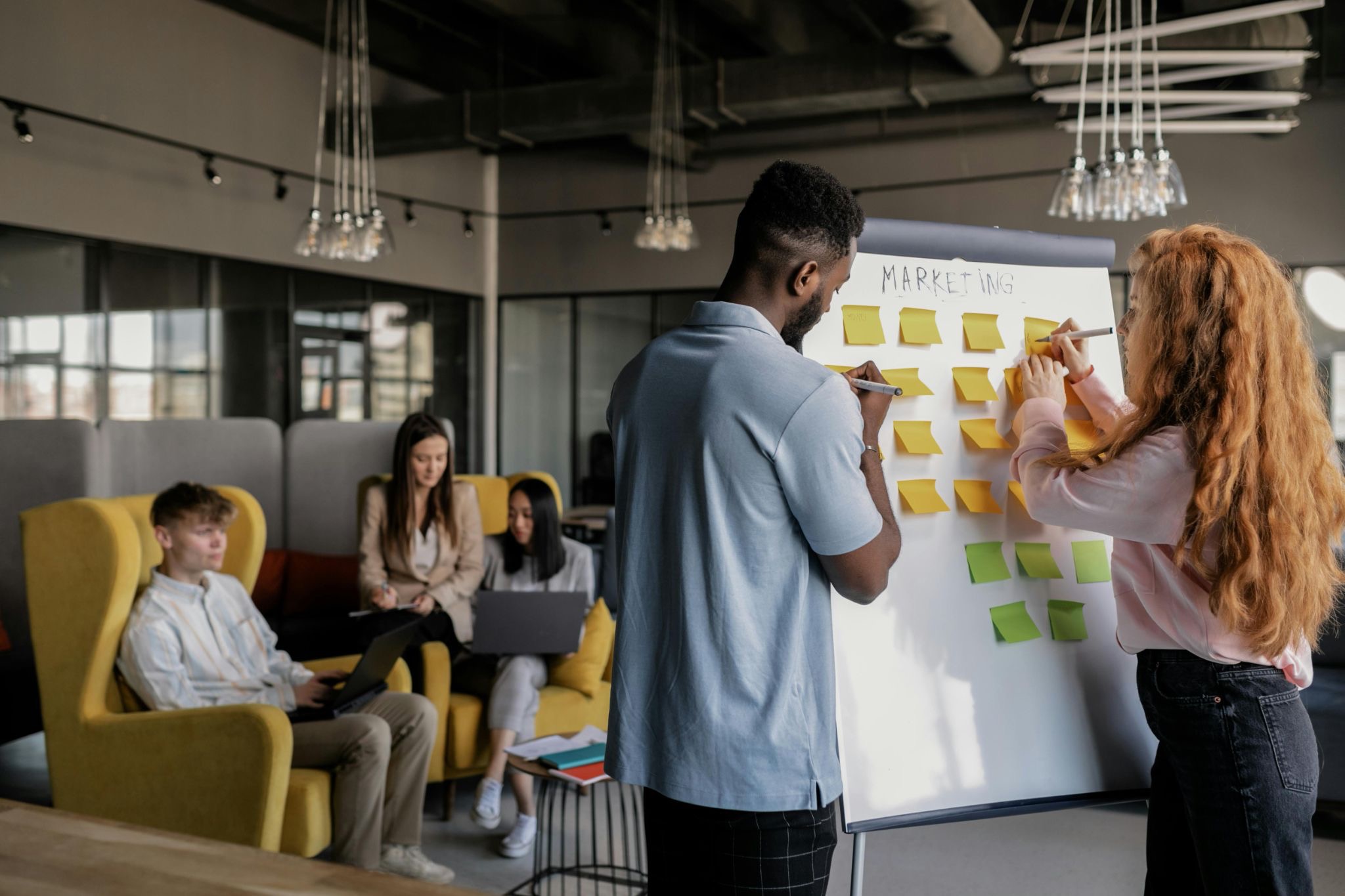 man and woman standing at whiteboard adding sticky notes to it. with people sitting in the background in an office