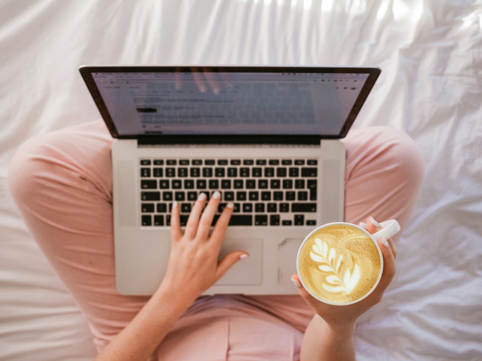 person using MacBook Pro and holding cappuccino on bed