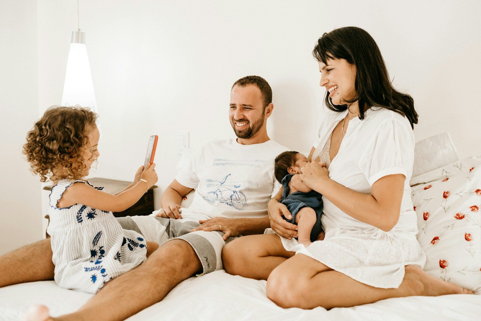 man isitting beside woman on bed holding a baby, with a young girl taking a photo of them with a phone