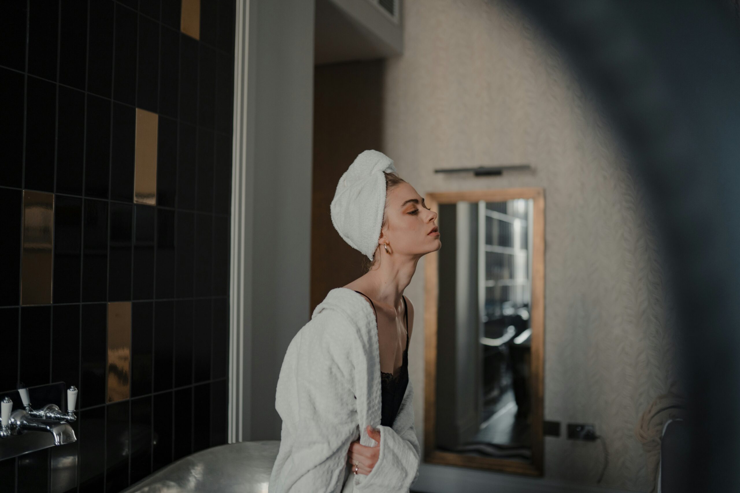 woman wearing white towel around head and white dressing gown, perched on the edge of bath