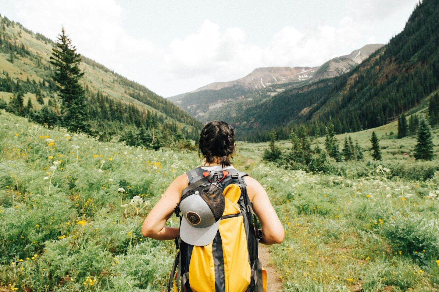 person carrying yellow and black backpack, walking on grassy field surrounded by hills