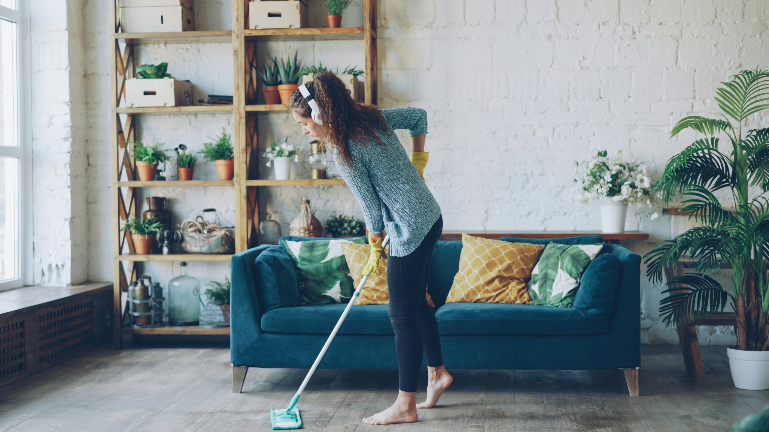 woman with dark curly hair wearing casual clothing is mopping floor at home and listening to music through headphones