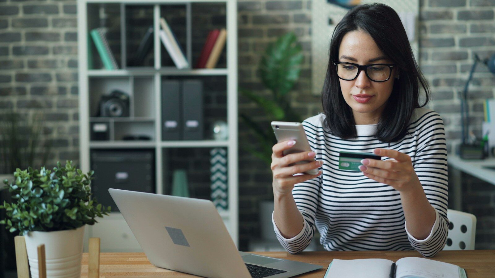 a woman sitting at a table looking at her phone with a bank card in other hand