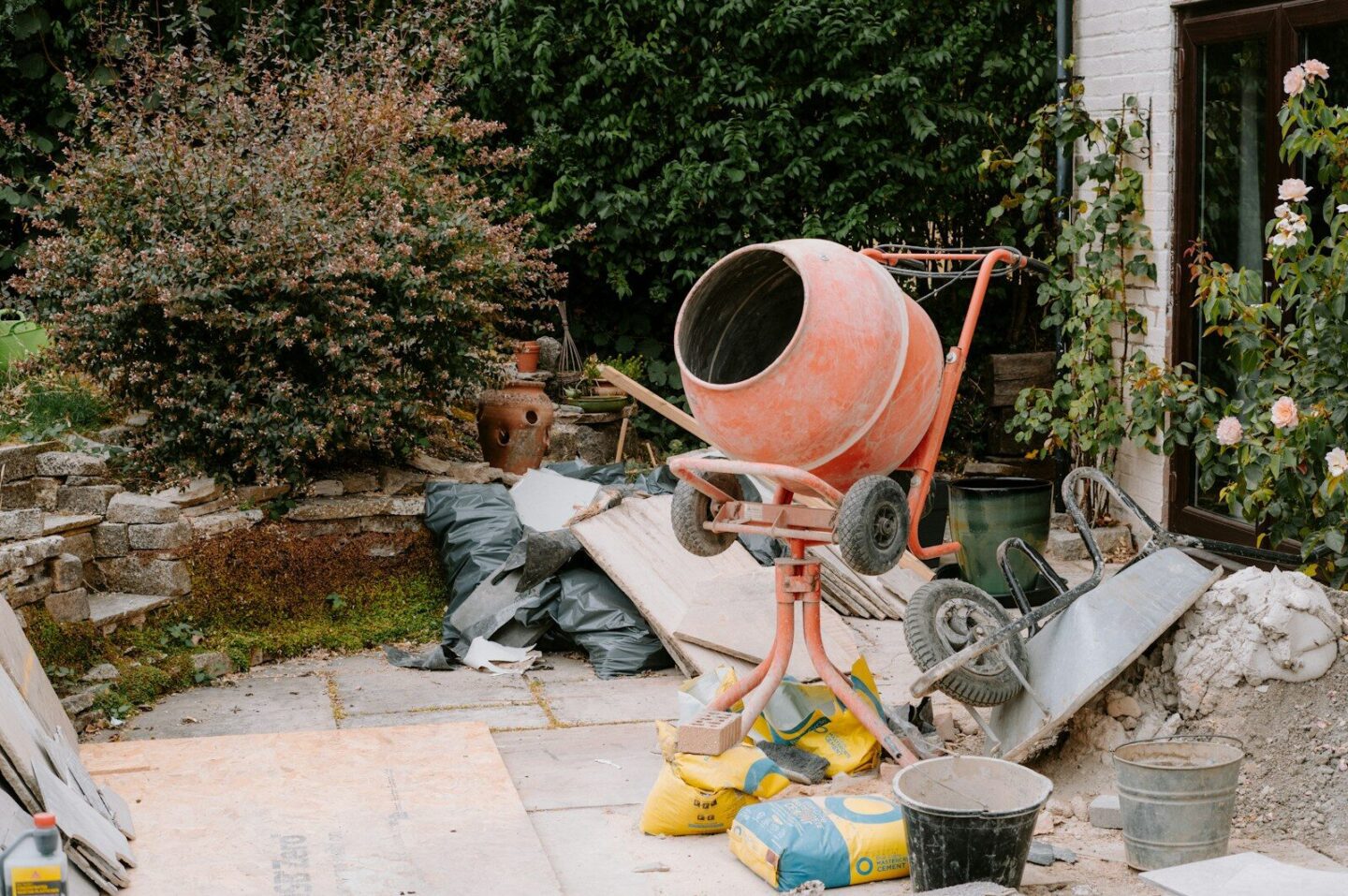 An orange cement mixer on a patio with construction materials and garden plants
