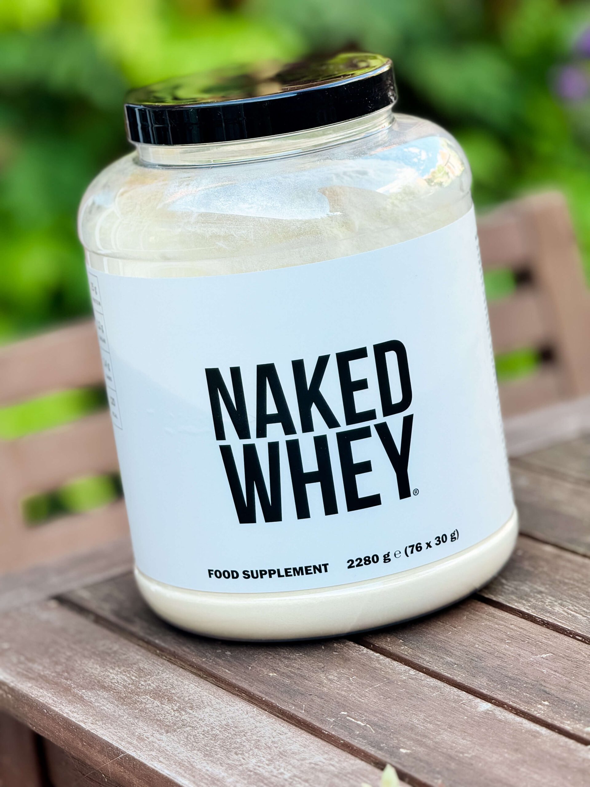 naked nutrition naked whey protein powder tub sitting on wooden table with greenery behind
