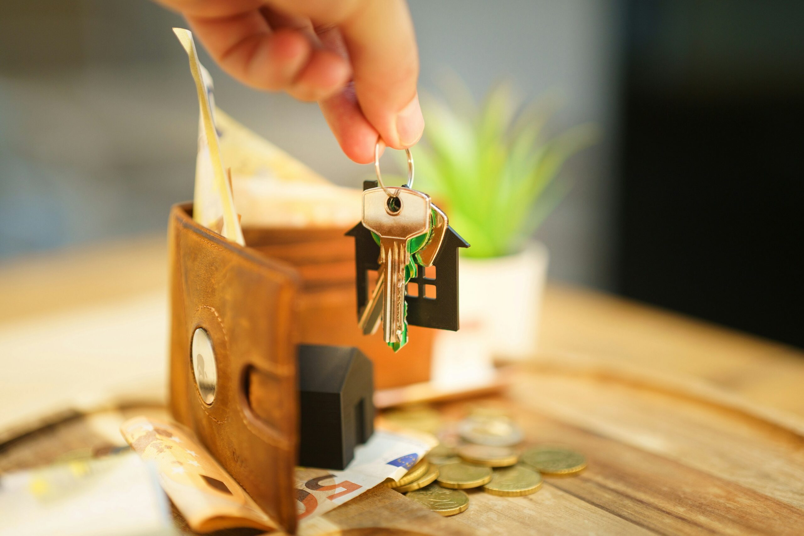 hand holding a set of keys above an open wallet, cash, and a black model house